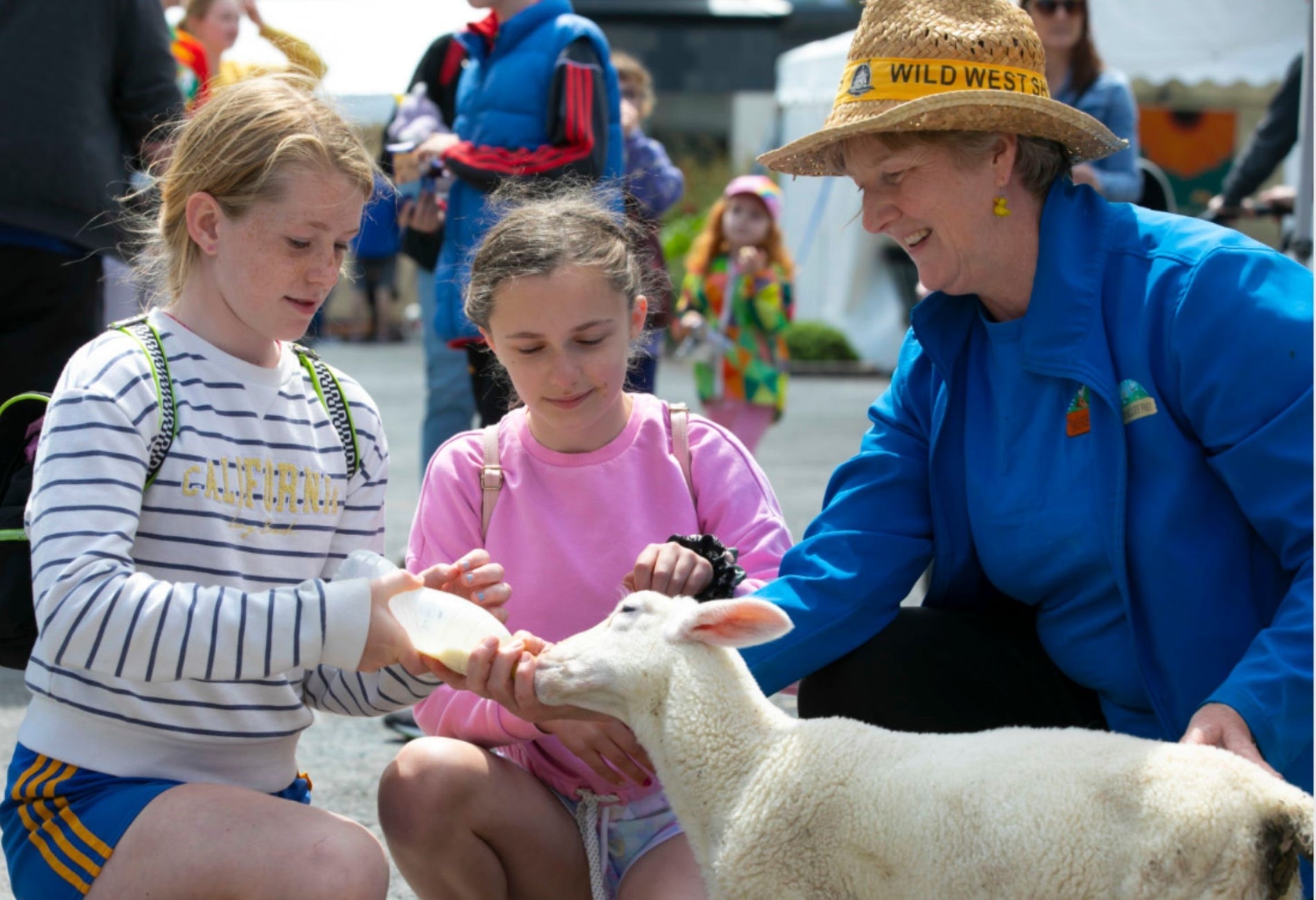 Three people crouched down with one holding a bottle feeding a lamb