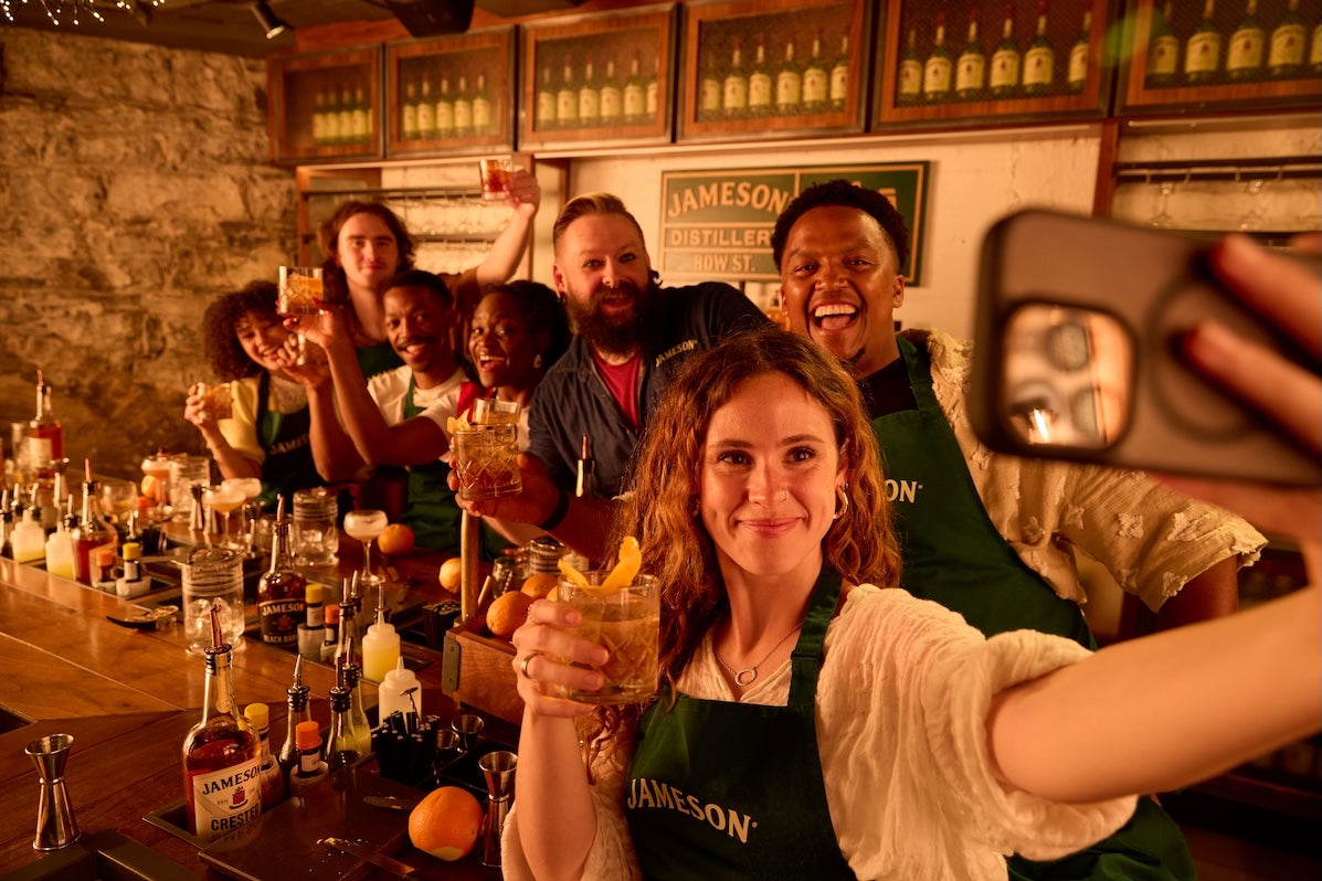 Group of  smiling people at a bar raising a glass with woman at the front taking a group selfie.