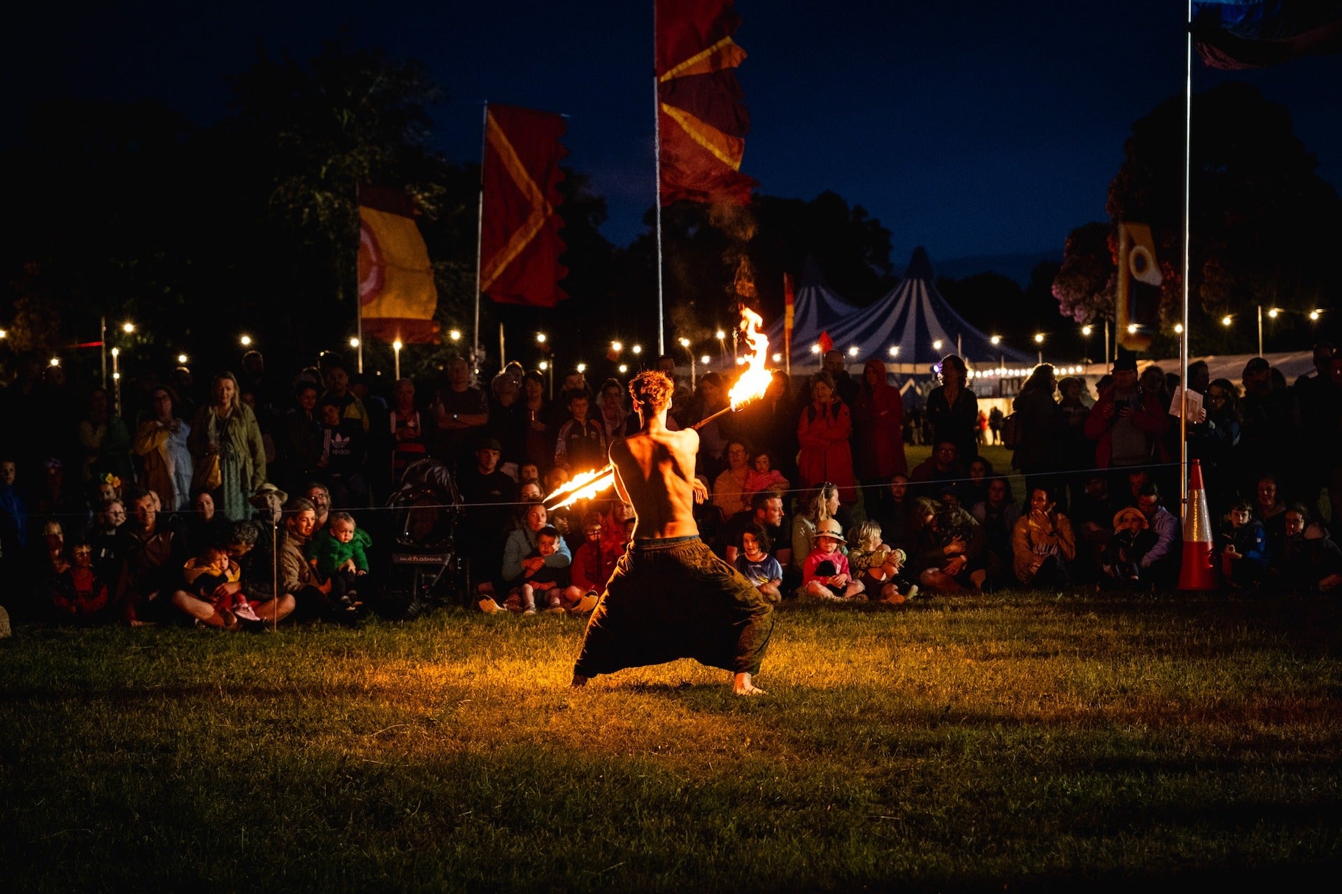 A night a crowd are watching a shirtless man who is moving with a fire stick.