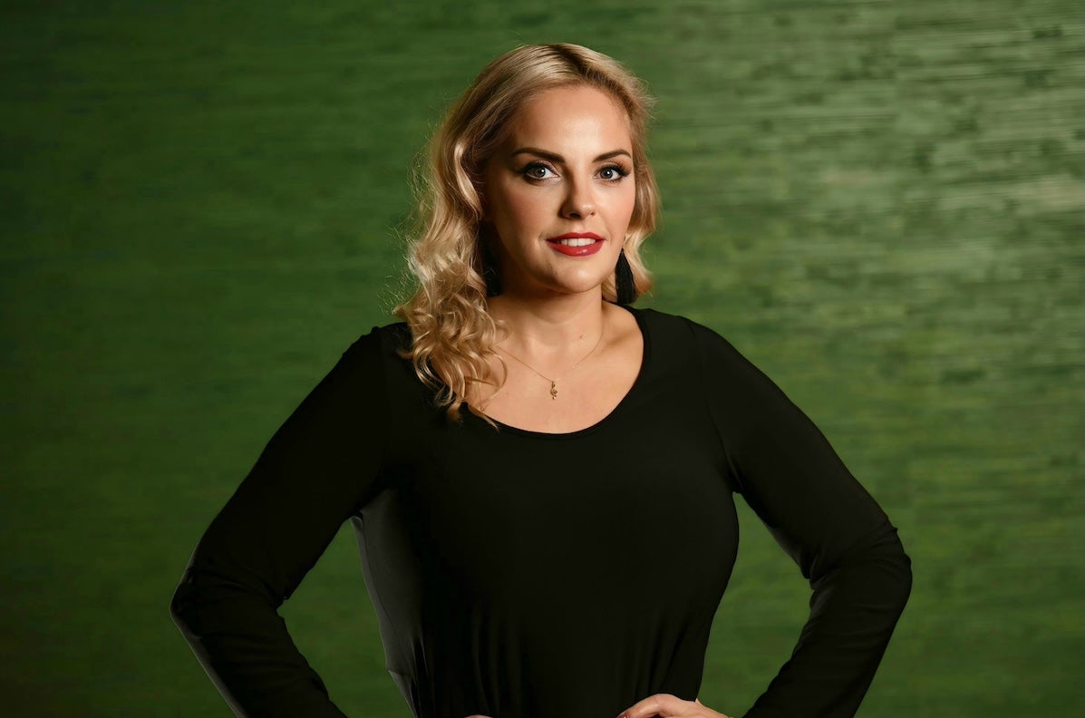 A gently smiling woman in black top with dark green background.