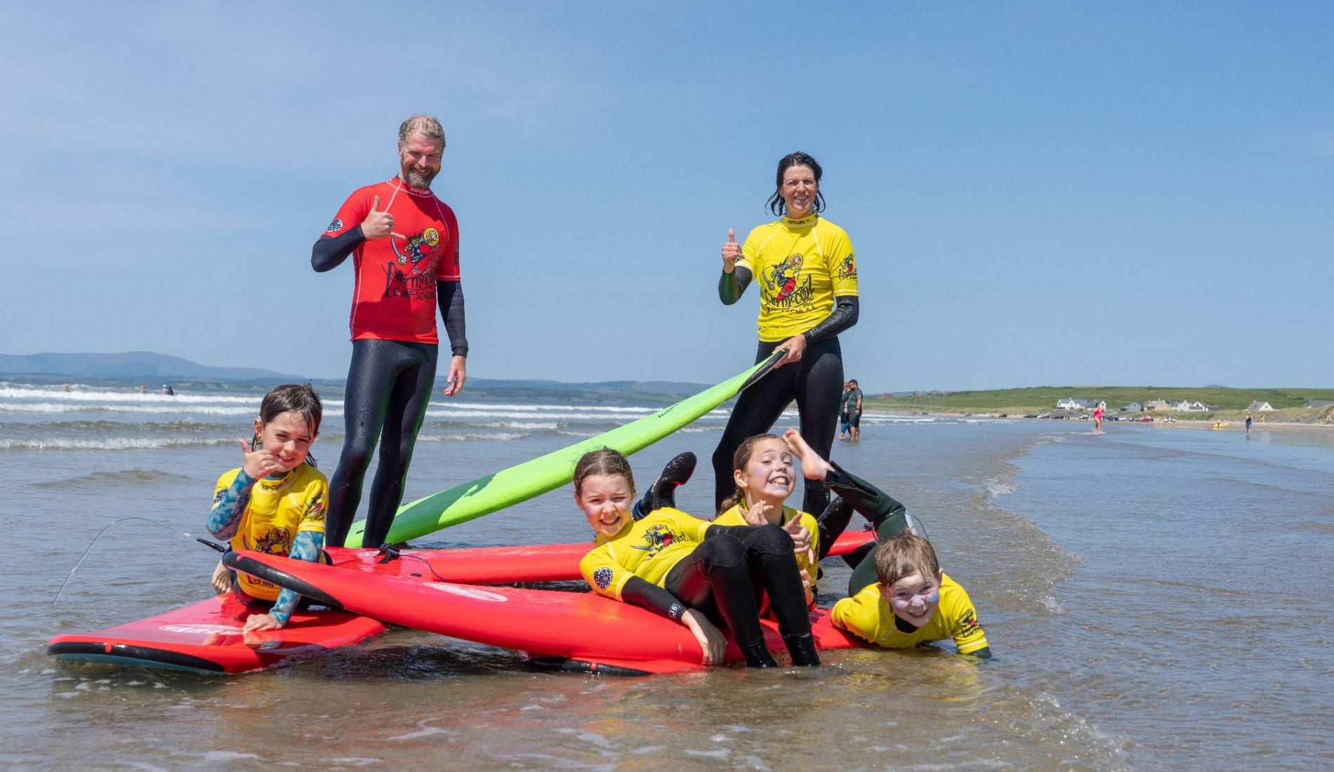 Surf instructors and children in wetsuits on the beach with red and green surfboards