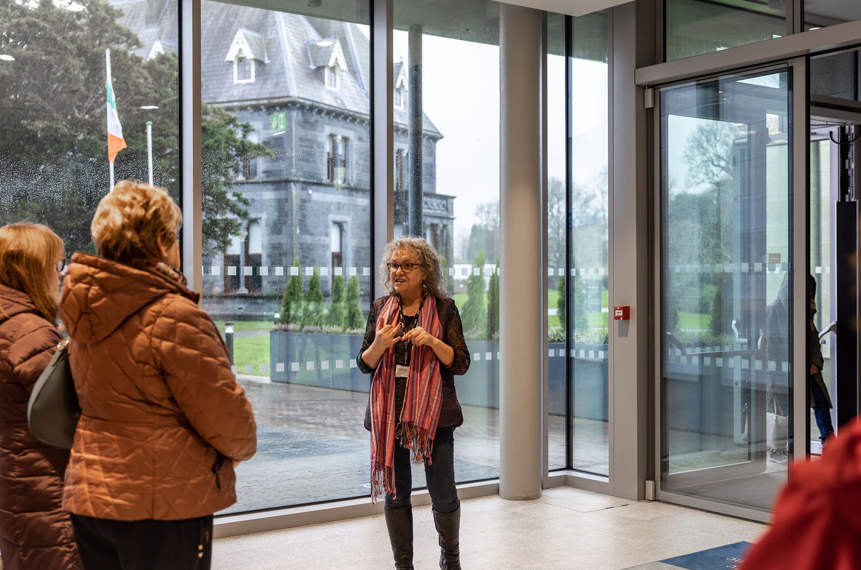 A woman tour guide in a glass lobby area is talking to small group with old, grey building visible through glass wall.
