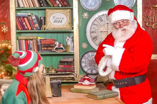 A santa is pointing a finger at a young elf seated at a wooden table with book shelf in background.