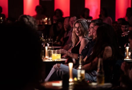 Audience members sitting at at table with drinks and laughing with red lights in the background