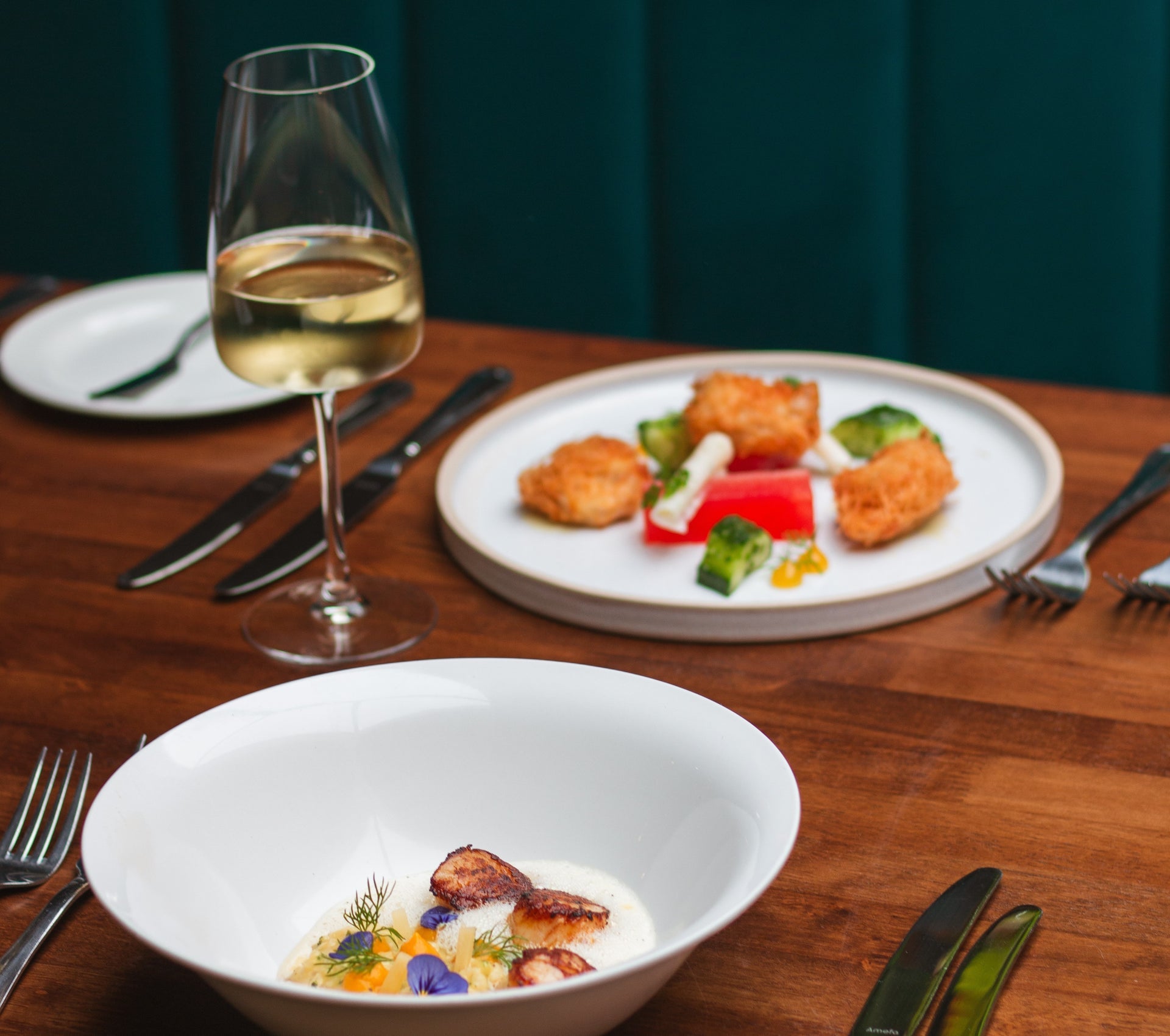 Table with two white plates of food a glass of wine and cutlery on it
