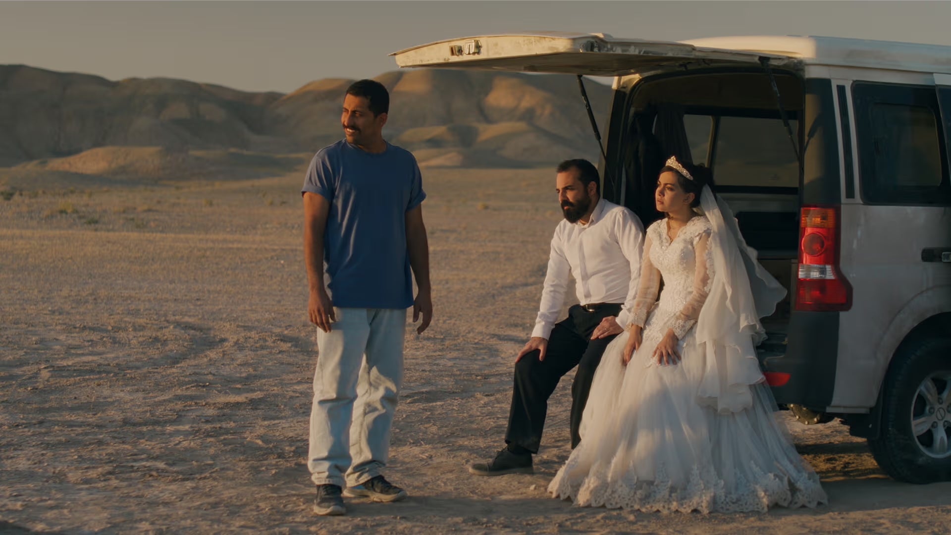 Two men and a woman in a wedding dress are waiting at the back of a vehicle in a desert.