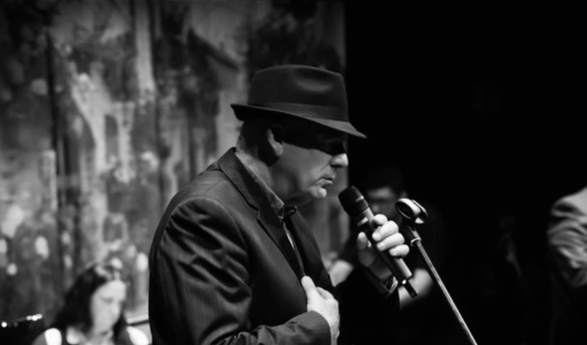 Black and white photo, side view of man in suit and hat singing into a mic.