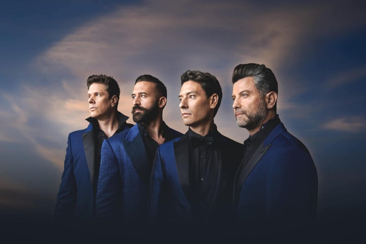 4 men in dark blue suits in a line all looking away to their right against murky sky.