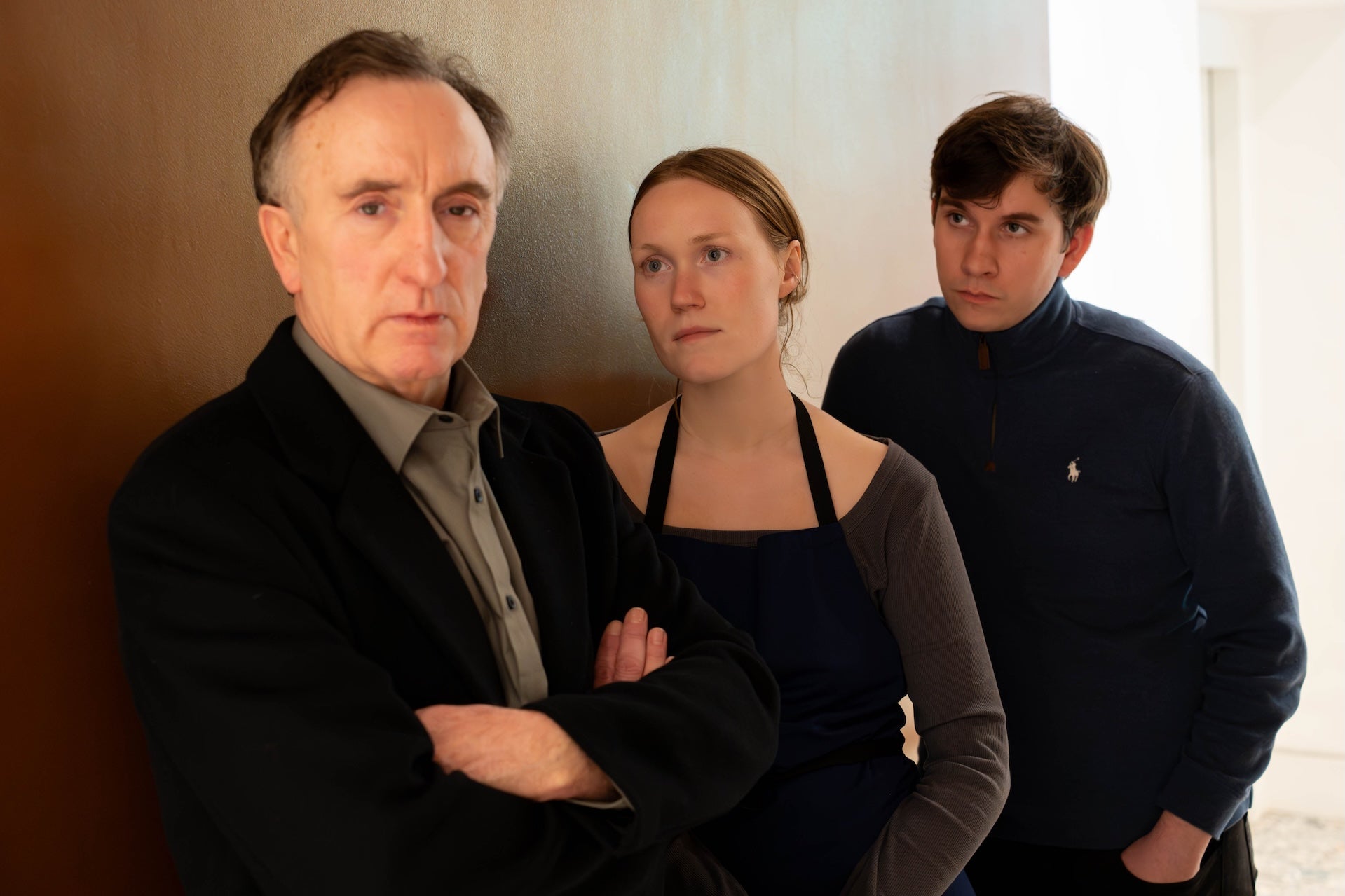 2 people are leaning against and internal wall looking at a man with crossed arms who is not looking at them.