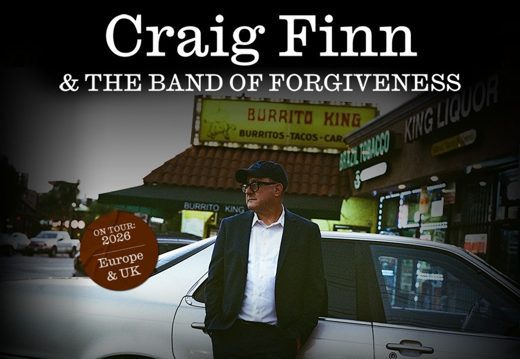 A man in jacket, white shirt and peaked cap is leaning against a silver car outside shops