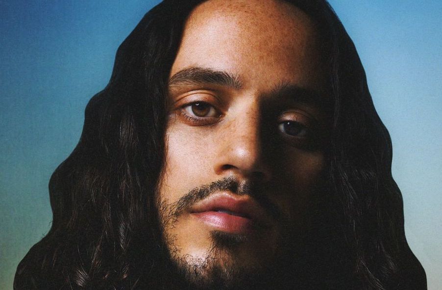 Close up head shot of man with short beard and long, wavy black hair against bluey background.