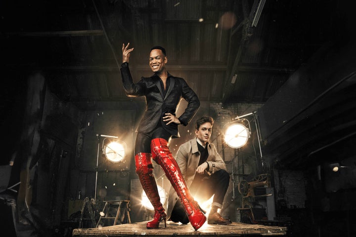 Kinky Boots The Musical, a smiling person in black wearing tall red shiny boots on a stage with another man crouching down behind him.