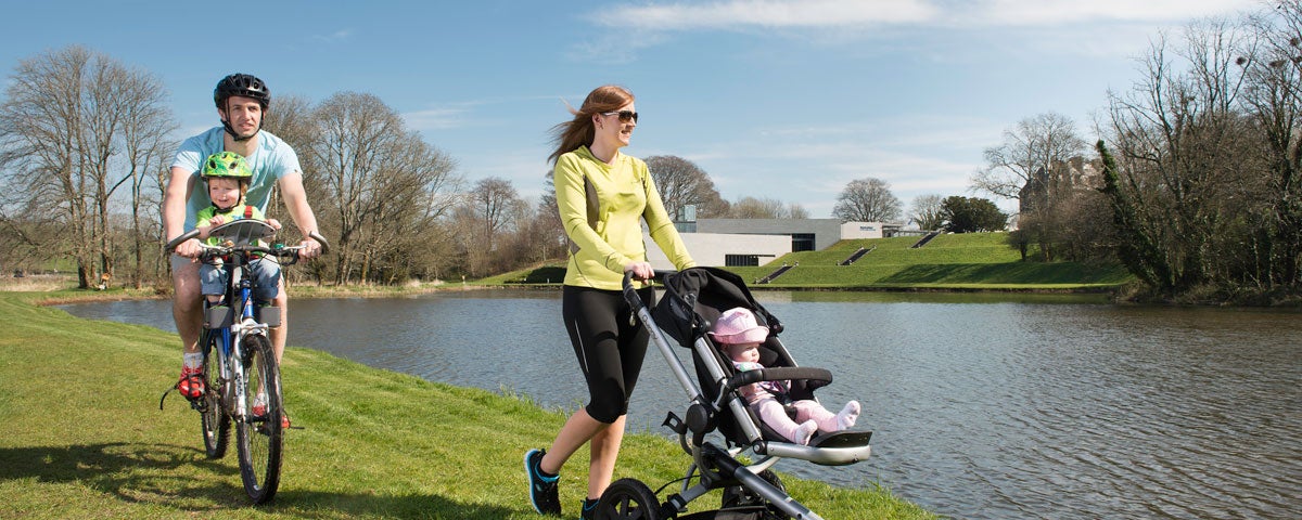Man on a bike with a child while a woman pushes a child on a buggy beside a river