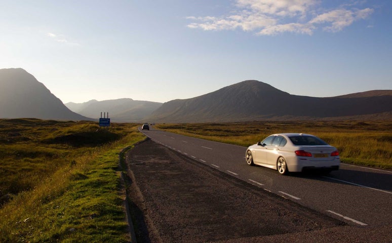 White car driving on a wide road towards mountains with green landscape on either side