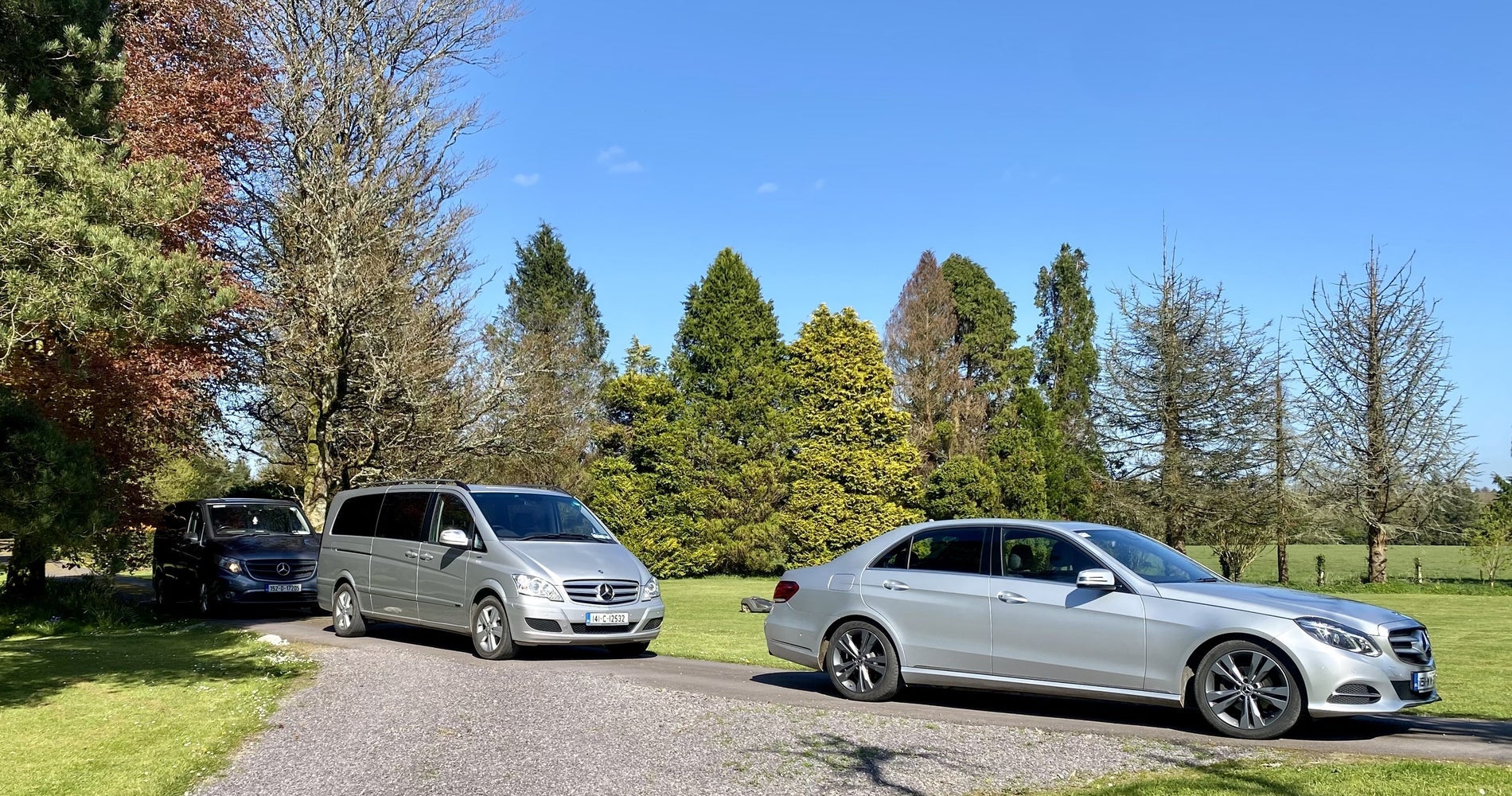 Three silver vehicles parked on a road in the middle of trees and a lawn