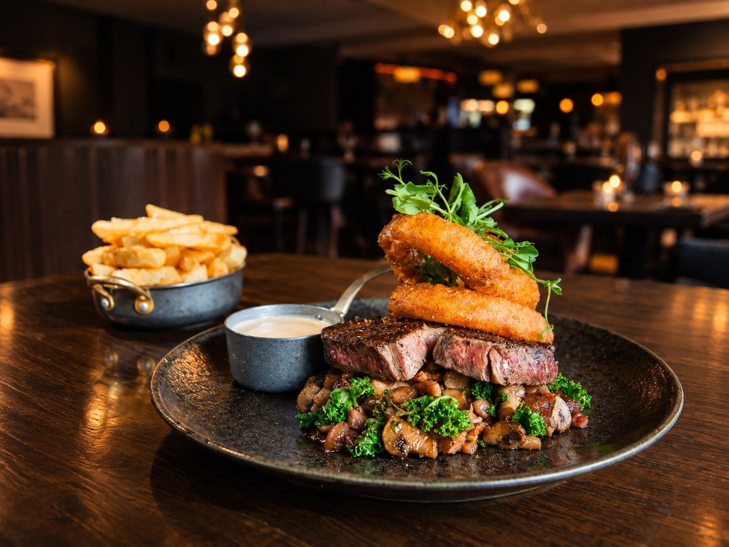 Steak with onion rings and a sauce on a plate with a portion of chips in the background