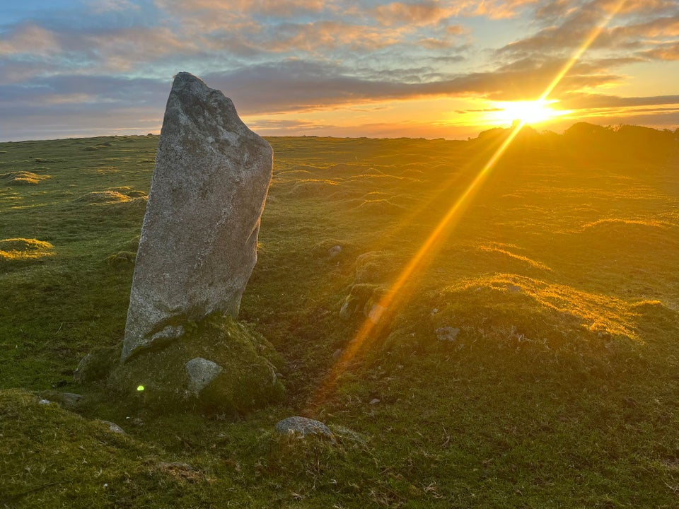 A granite standing stone, with a sunrise breaking through cloud behind it