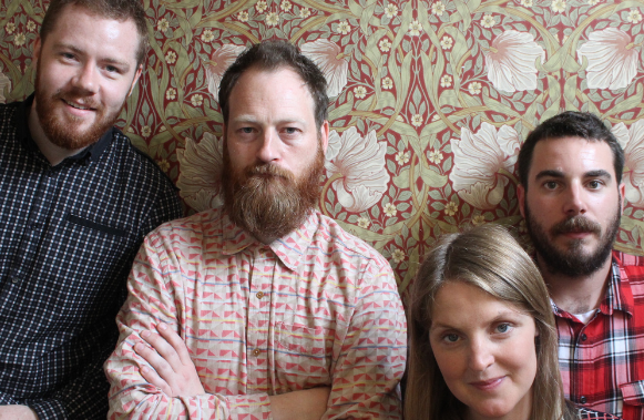 The 4 members of Roamer standing against a wall of ornate wallpaper.