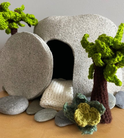 A stone tomb, open, with knitted trees and smaller stones surrounding it.