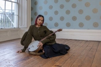 A woman with stringed instrument seated on a bare wooden floor in an empty room