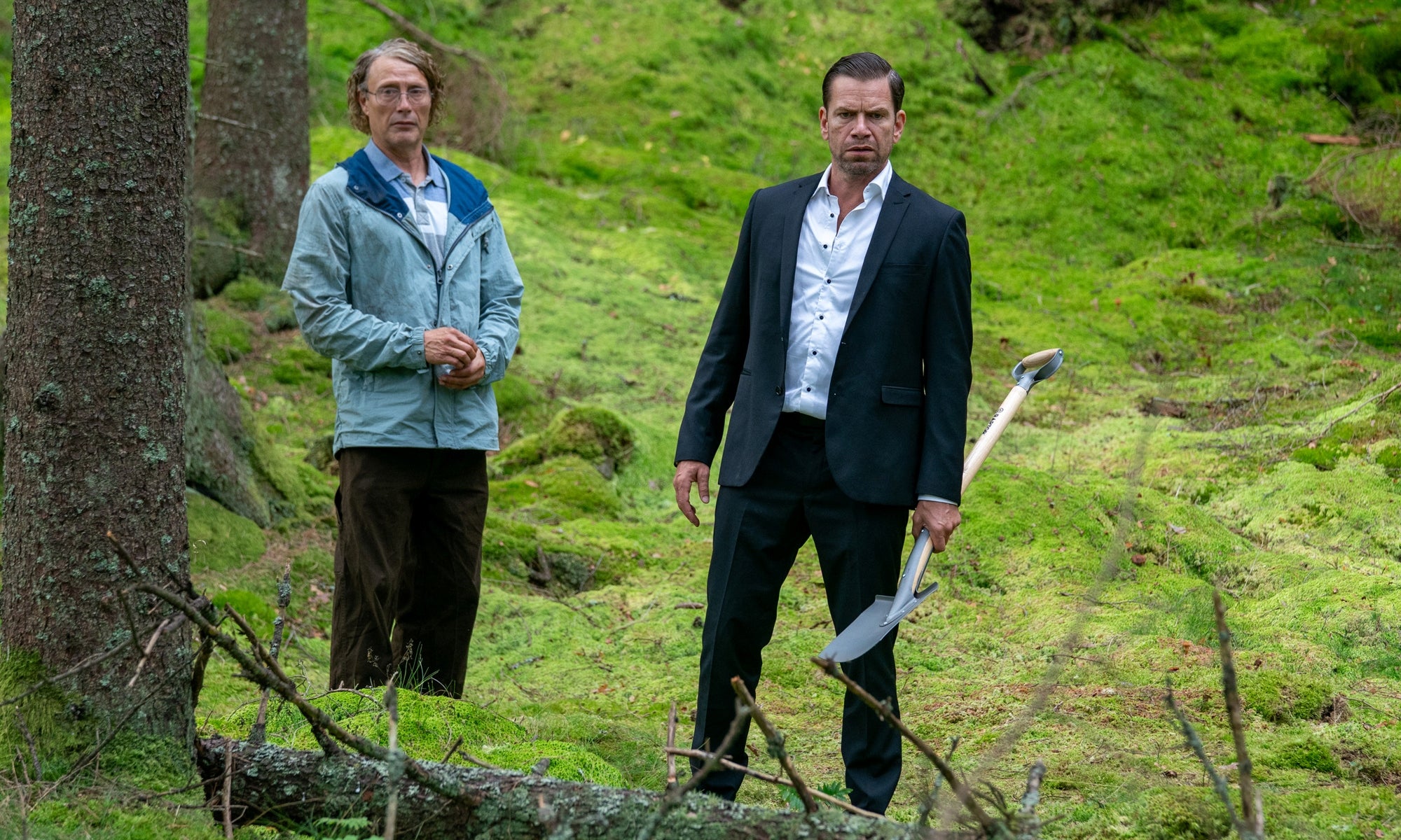 A man in a crumpled raincoat and a man in a suit are in a forest.