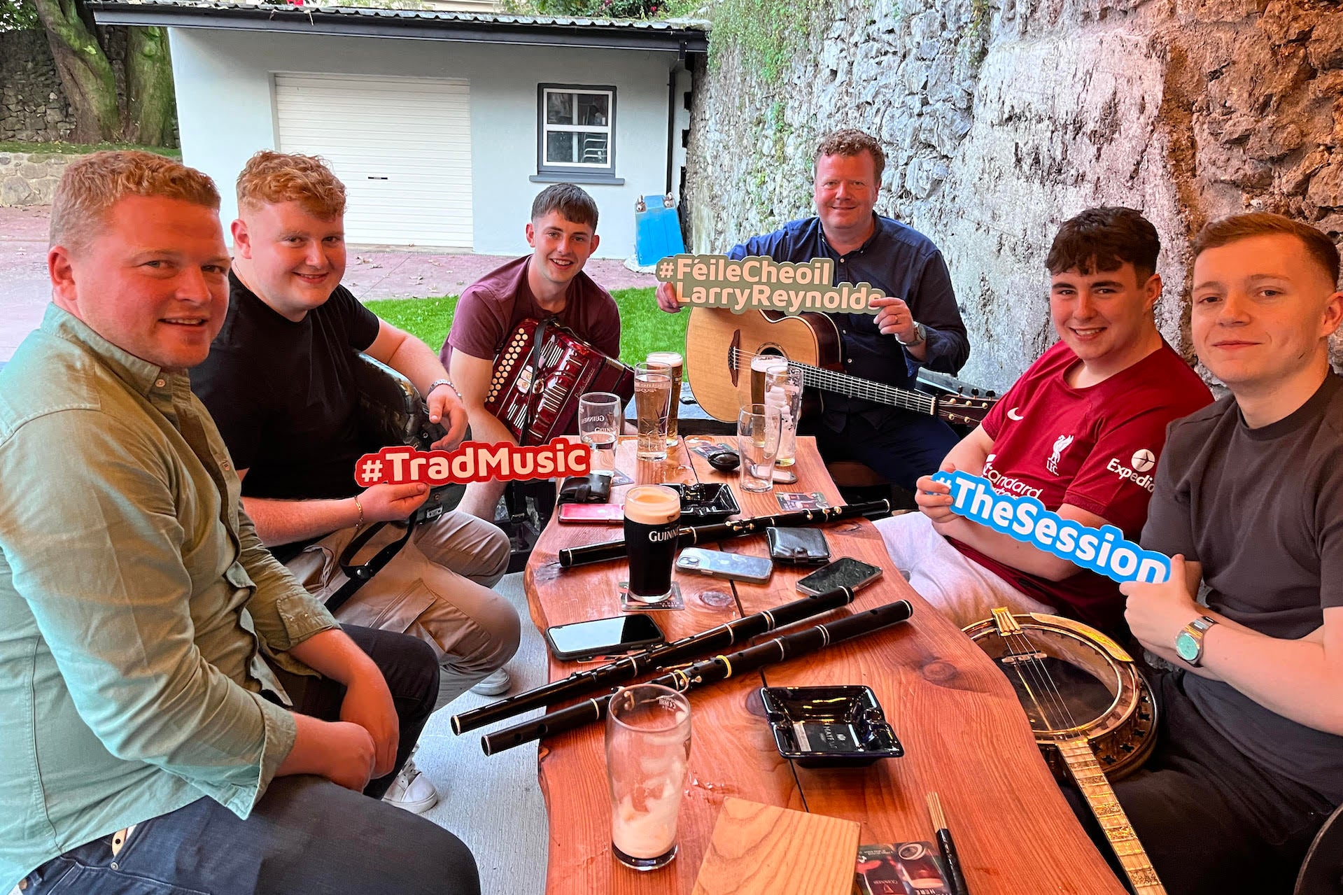 Live Music Session with top musicians in Beer Garden, 6 seated around a table outdoors with musical instruments