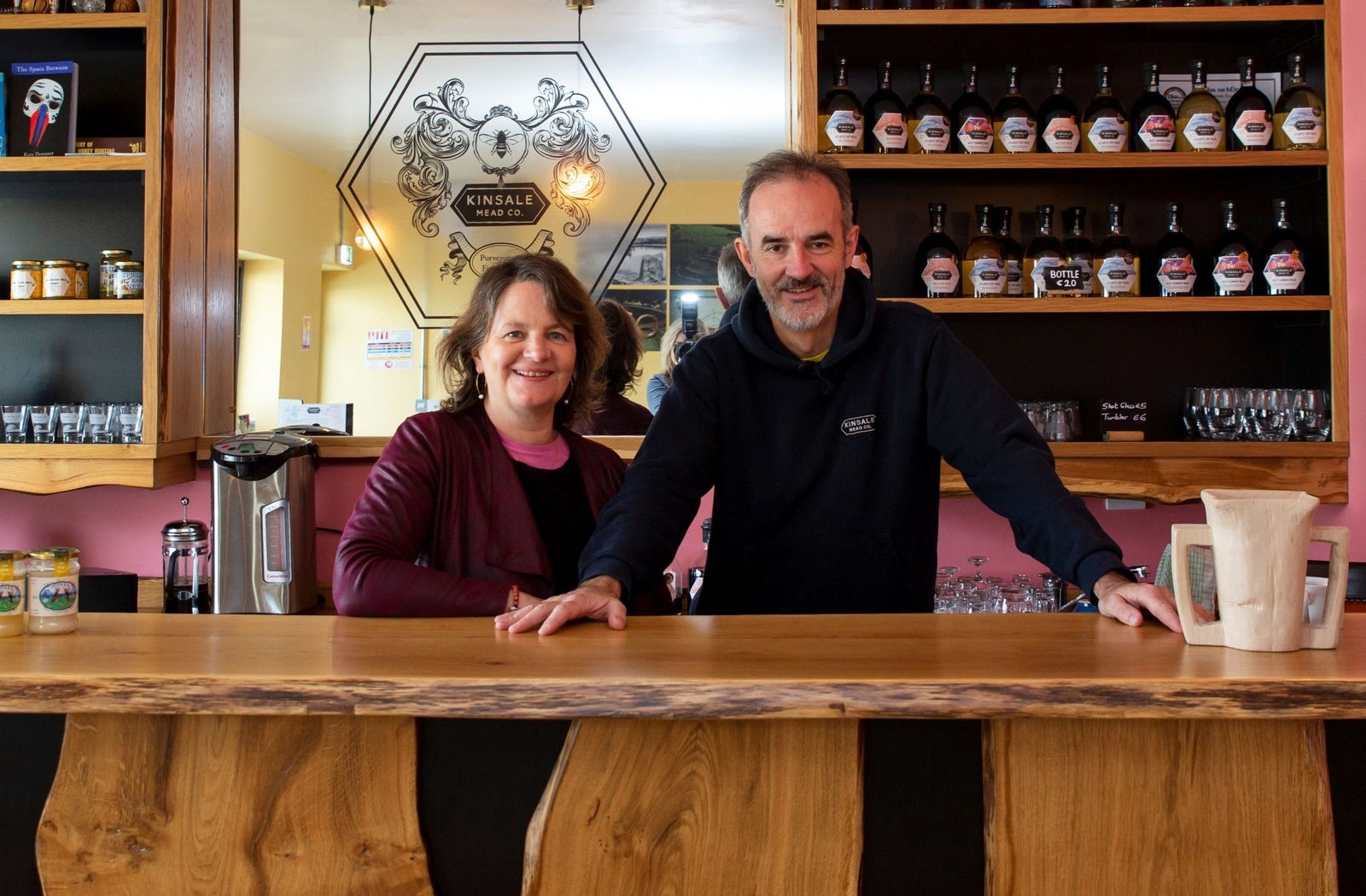 Two people standing at a wooden counter with displays of bottles on shelves behind them