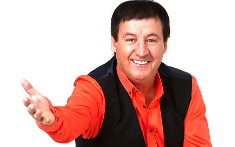 A smiling man in redshirt and black open waistcoat holding his right hand foreward.