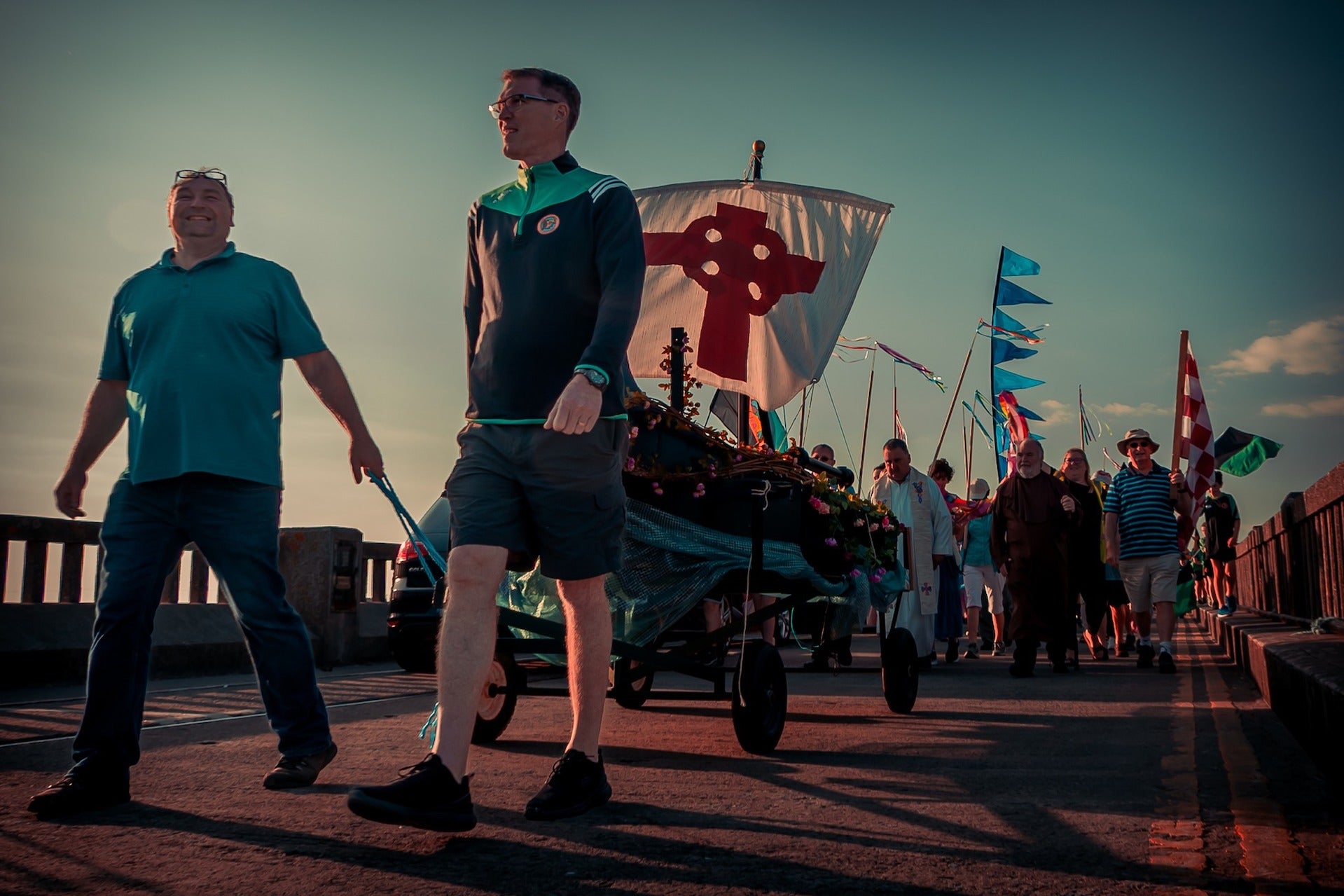 St Brendan the Navigator Community Festival 2026, at dusk, 2 men are pulling a small trolley in a parade with flags.