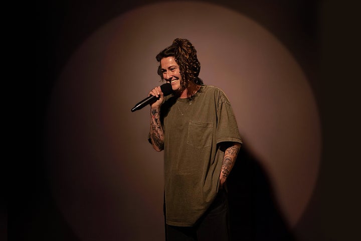 A smiling woman is holding a mic up to her mouth, against dark brown background.