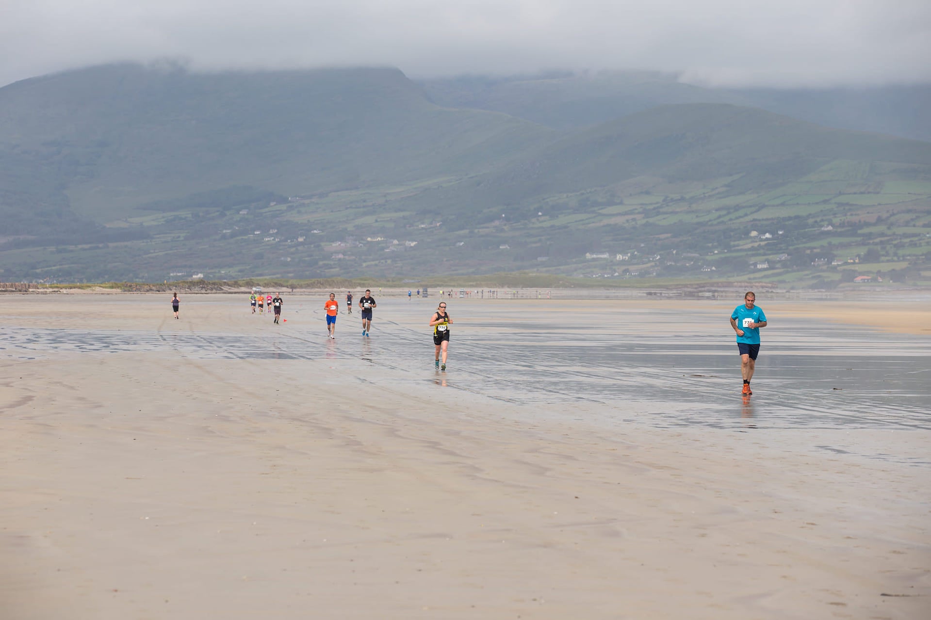 View of a large beach with people running along it, against background of hills