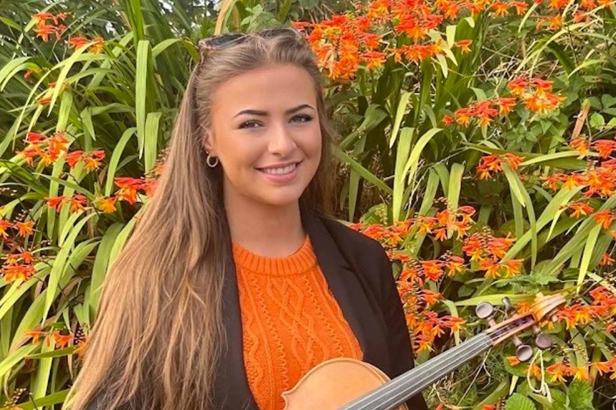 A smiling young woman holding a fiddle with large flowering plant in background.