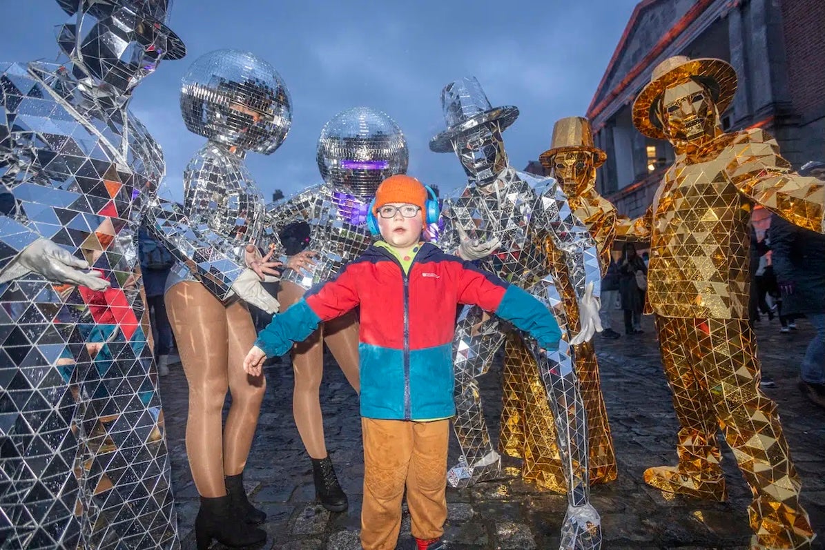 A child outdoors in coat and hat standing with group of 6 people in silver or gold mosaic covered costumes with faces hidden by headwear.