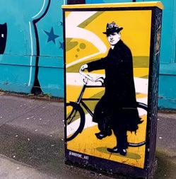 Street art of Michael Collins wearing a long black coat and hat with his bicycle on a yellow background