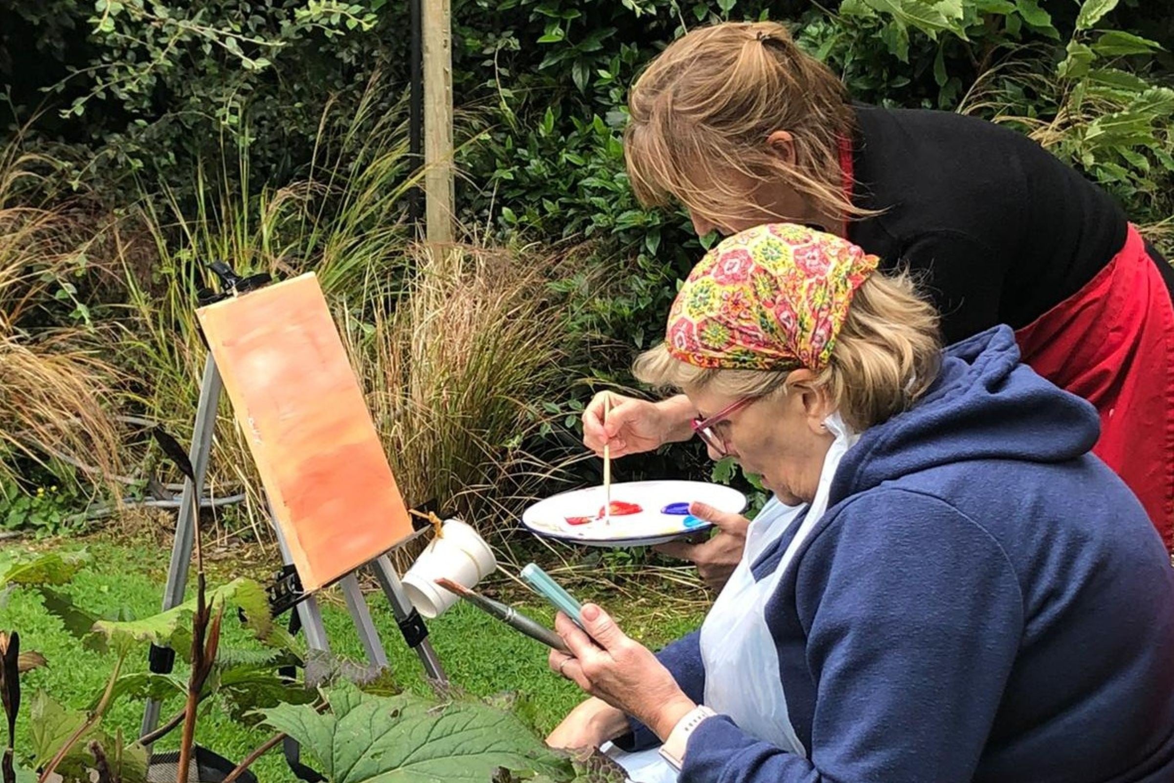 An artist sitting outdoors at an easel while another artist mixes paint on a palette
