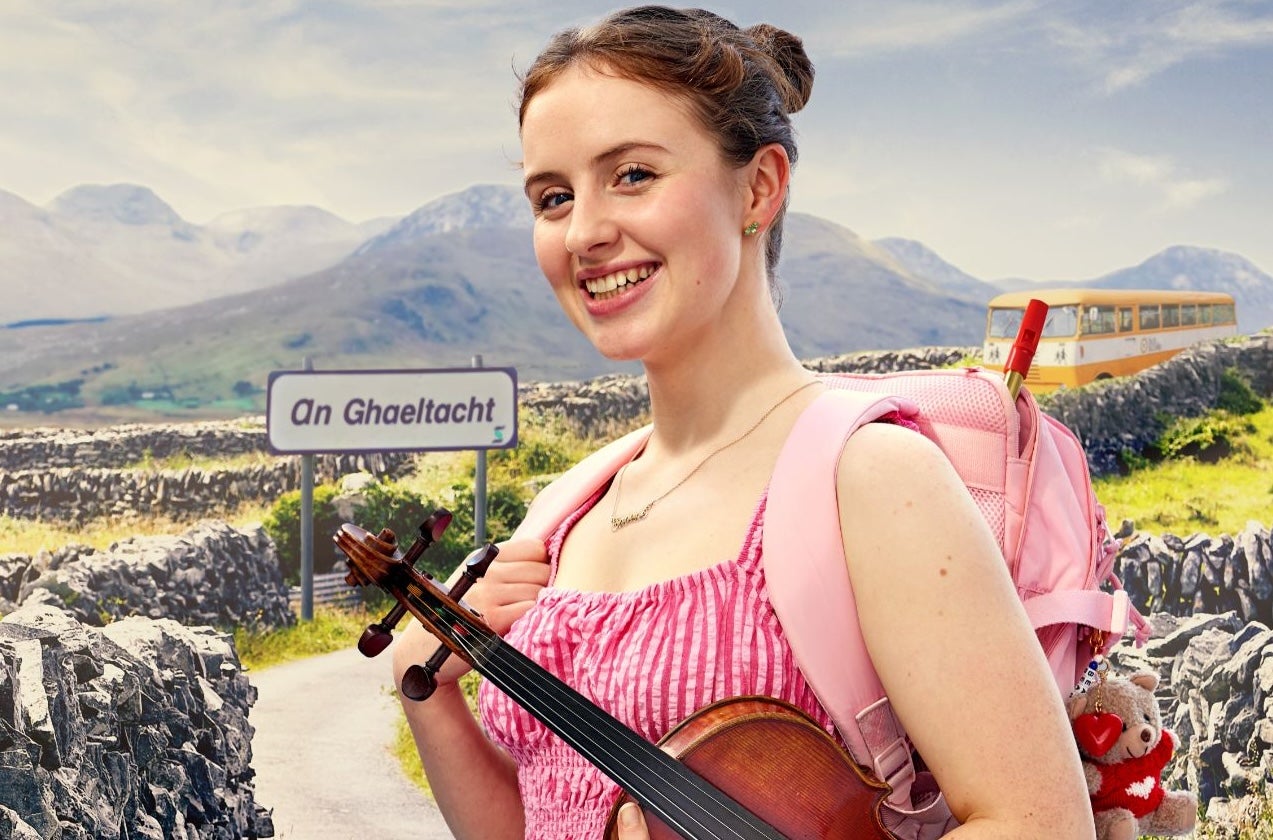 Ciara Cox holding a violin standing in front of a Gaeltacht sign and bus with stone walls and mountains in the background