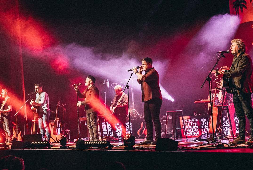 Looking up at a stage where a large group of musicians are playing and a man in the middle singing into a mic on a stand, lit with red and purple lights.