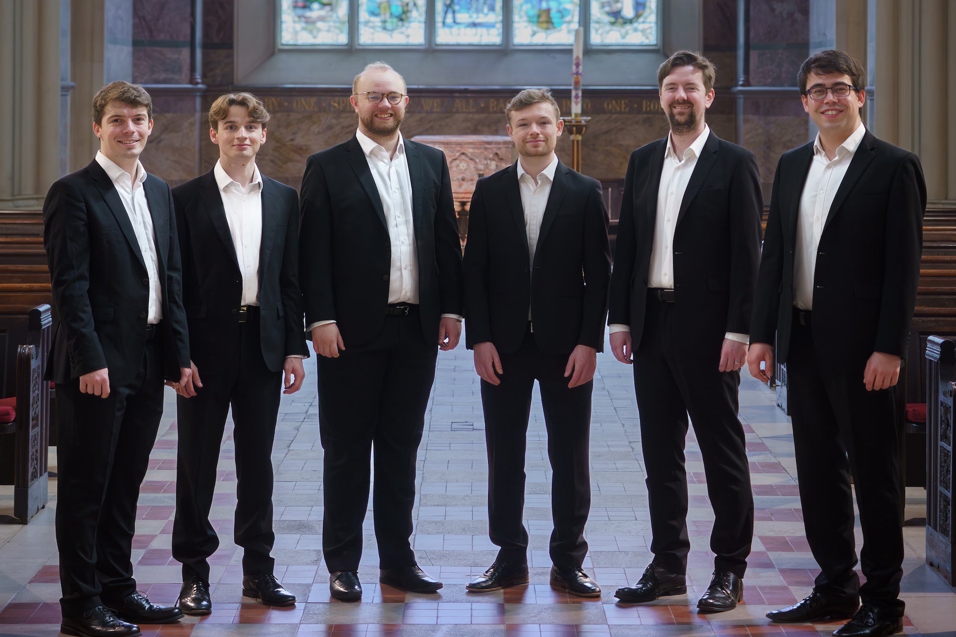 6 smiling men in dark suits and white shirts a in a line in a wide church aisle