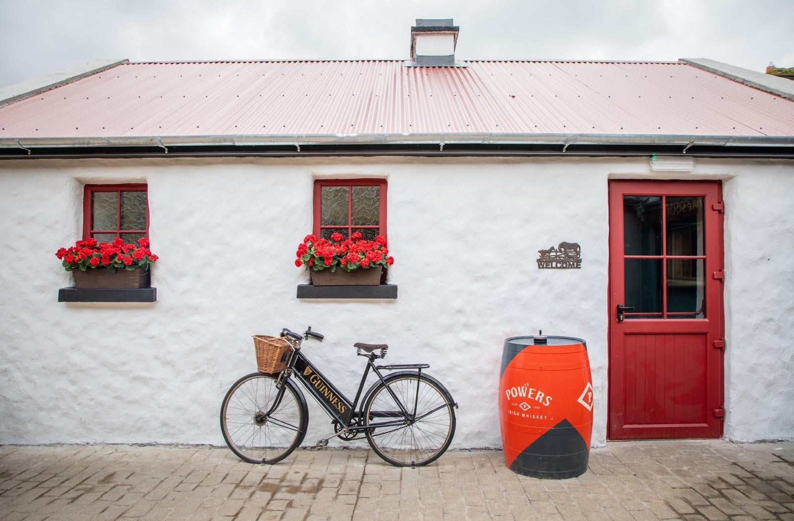 White cottage with red windows and door and a bike and barrel outside