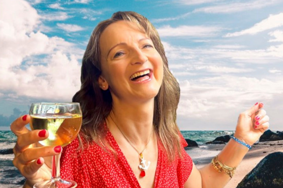 A laughing woman holding a glass of wine with beach in the background.