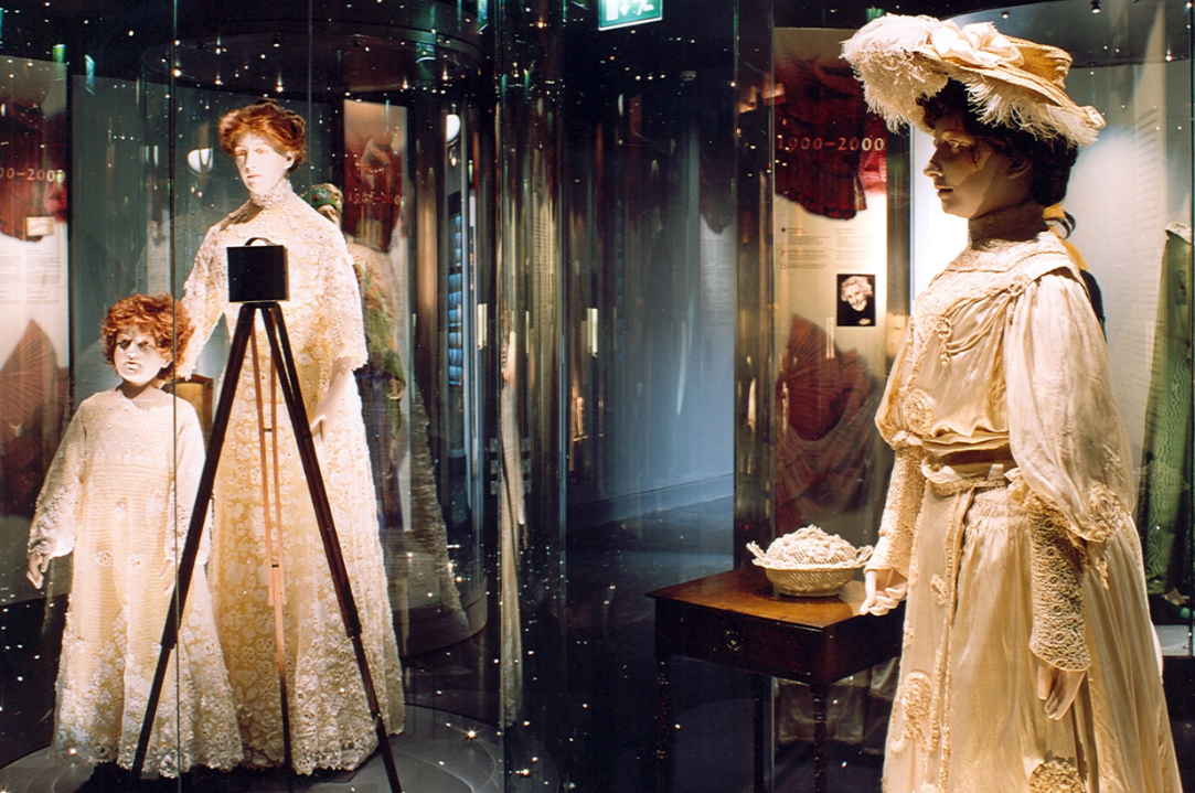 Display cabinets containing life size models wearing old clothing in beige colours in dimly lit room.