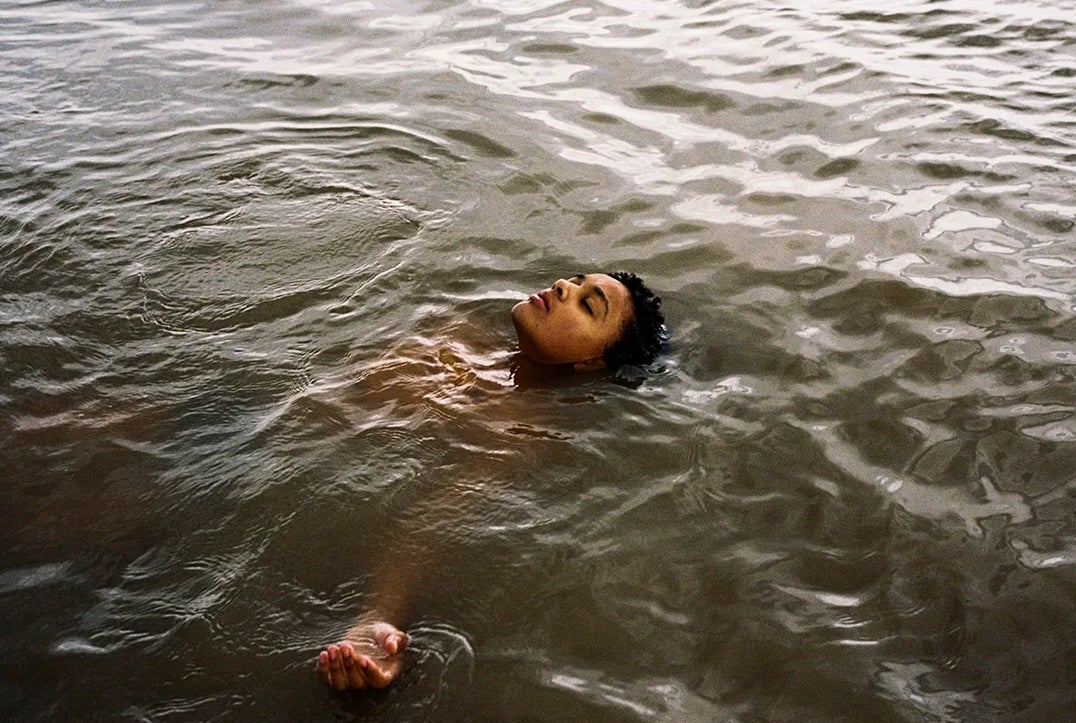 A young person is floating on their back in grey water with head and one hand visible above the water.