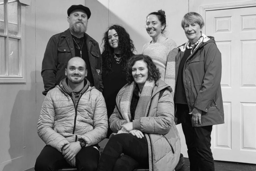 A group of smiling people with 2 seated, the rested standing, some wearing coats indoors.
