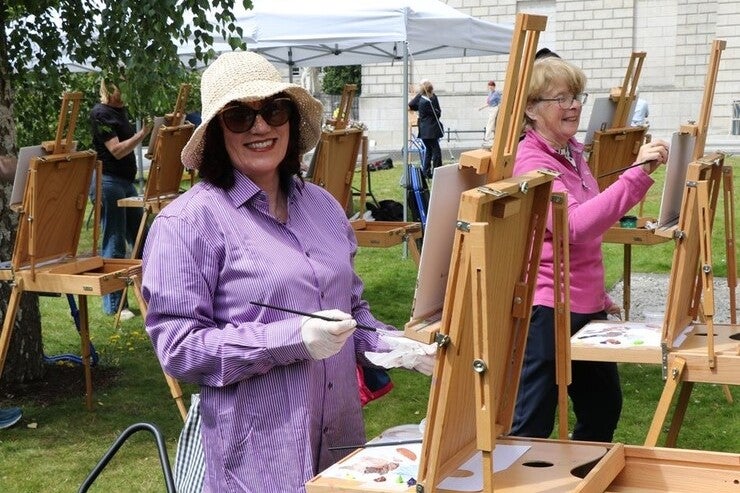 Easels set up outdoors with smiling woman in a hat painting