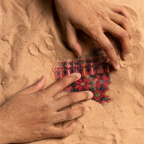 Looking down at a pair of hands digging out a small square of red and black patterned cloth from sand.