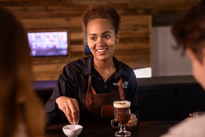 A smiling person in apron with 2 drinks in front on a wooden counter.