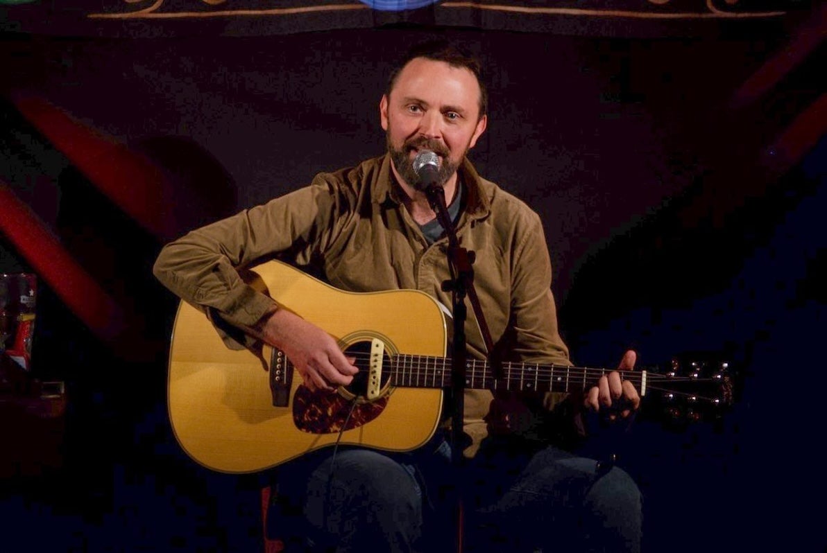 A man seated playing a guitar, singing into a mic on a stand