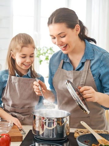 A woman and young girl wearing identical aprons are smiling, looking down at steaming saucepan on a stove
