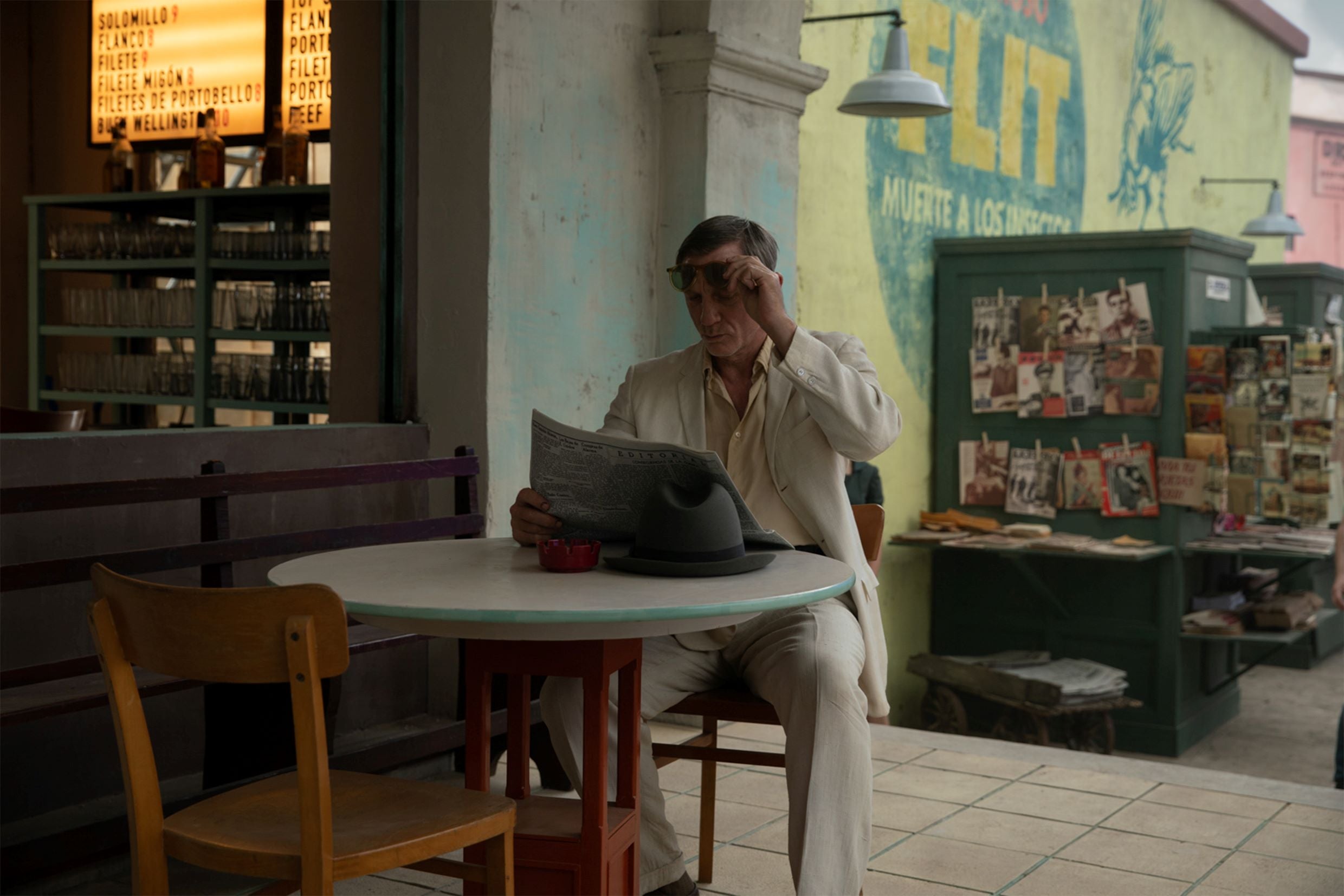 A late middle-aged man in a white suit is reading a newspaper at a cafe table.