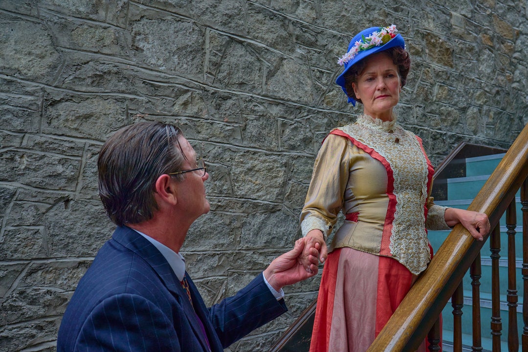A woman in early 1900 century dress and hat is on staircase, holding out hand to man at the bottom of the stairs.